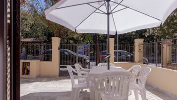 Een terras buiten met een tafel en stoelen onder een witte parasol, omringd door een metalen hek en geparkeerde auto's.