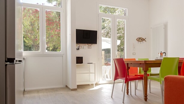 Una habitació amb taula de menjador, cadires, televisió penjada a la paret i una finestra que mostra vistes a arbres.