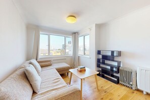 Ein modernes Wohnzimmer mit einer Couch, einem kleinen Tisch, einer Heizung und Blick auf die Stadt durch große Fenster.