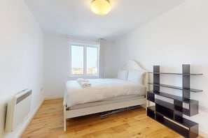 Ein Einzelbett mit weißer Bettwäsche, ein Heizkörper, ein Fenster mit Vorhängen und eine schwarze Regaleinheit an der Wand.