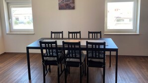 Uma área de refeições com mesa, cadeiras, duas janelas e um quadro na parede.