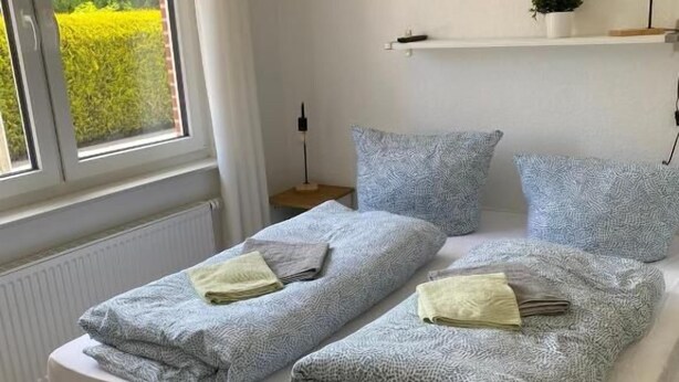 A double bed with a white mattress and grey bedding, a bedside table with a lamp, a window with curtains, and a shelf with a plant. - Fischerspfad (Norden)