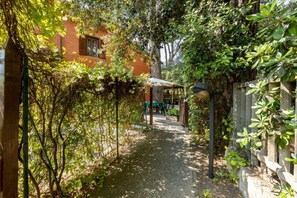 A pathway leading to a covered outdoor area with a table and chairs, surrounded by dense foliage and a brick building. - GuestHost - La Pineta Charming Home in Tirrenia (Tirrenia)