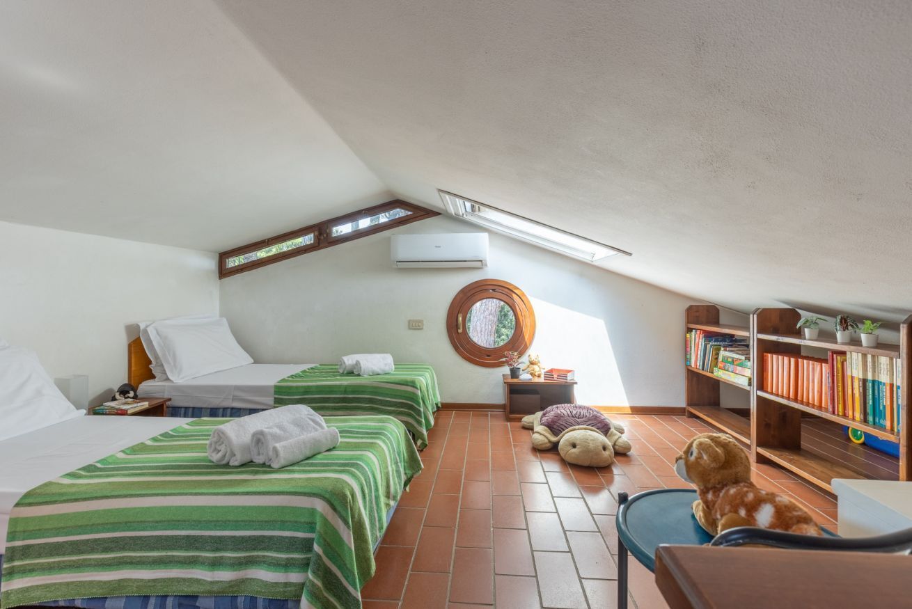 A room with two beds, a small table, a chair, a bookshelf, and a window with a view.
