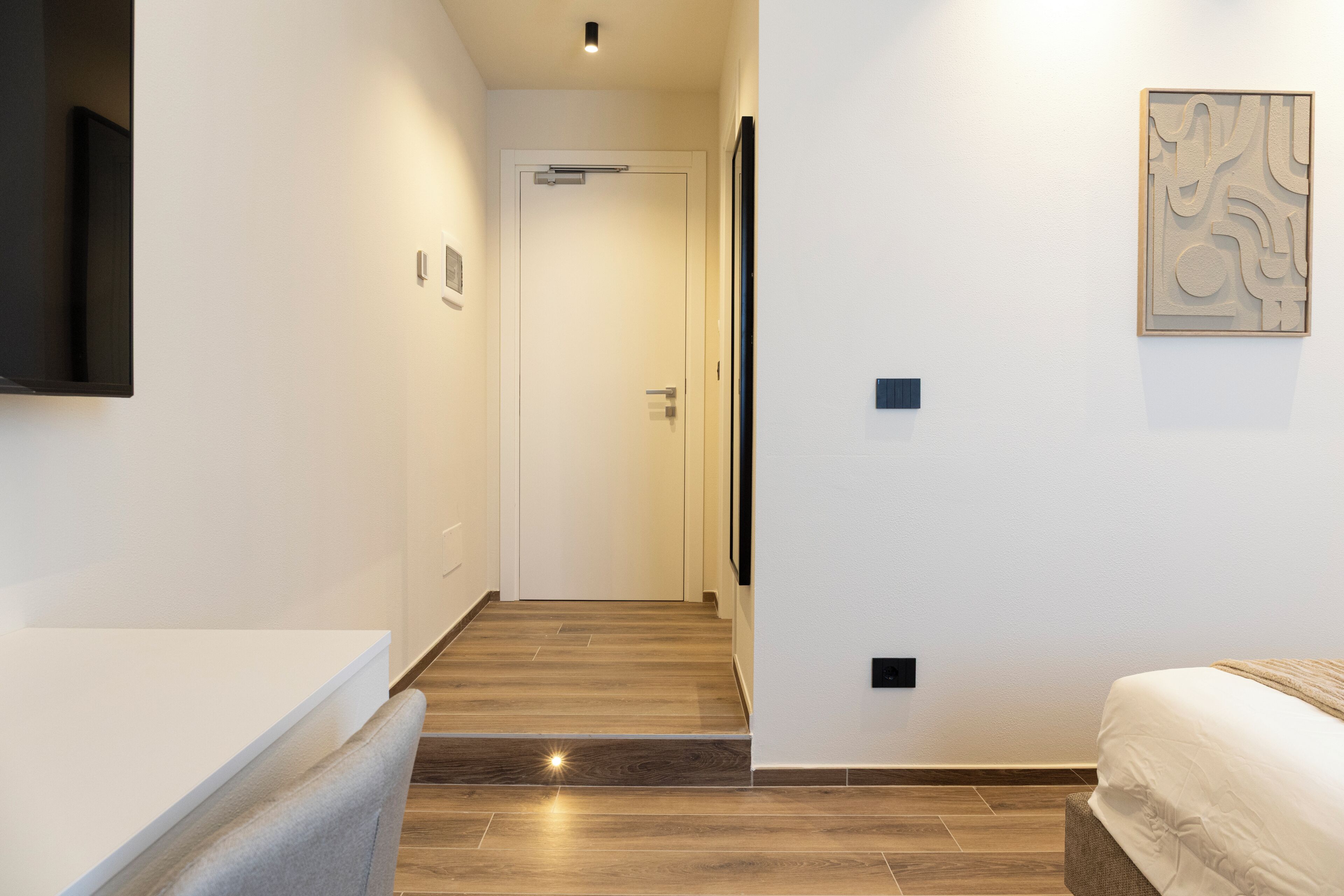 Habitación de hotel con piso de madera, cama, mesita de noche, televisor instalado en la pared y una puerta que da a otra habitación.