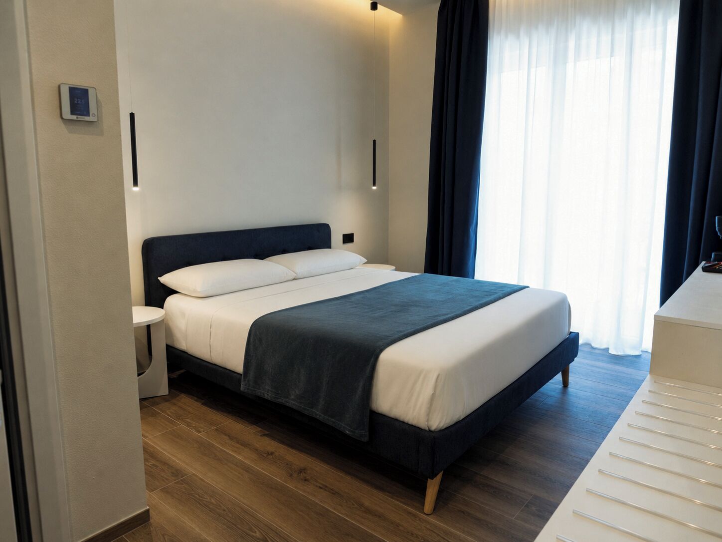 A modern hotel room with a bed, bedside table, and a view of the outdoors through a window.