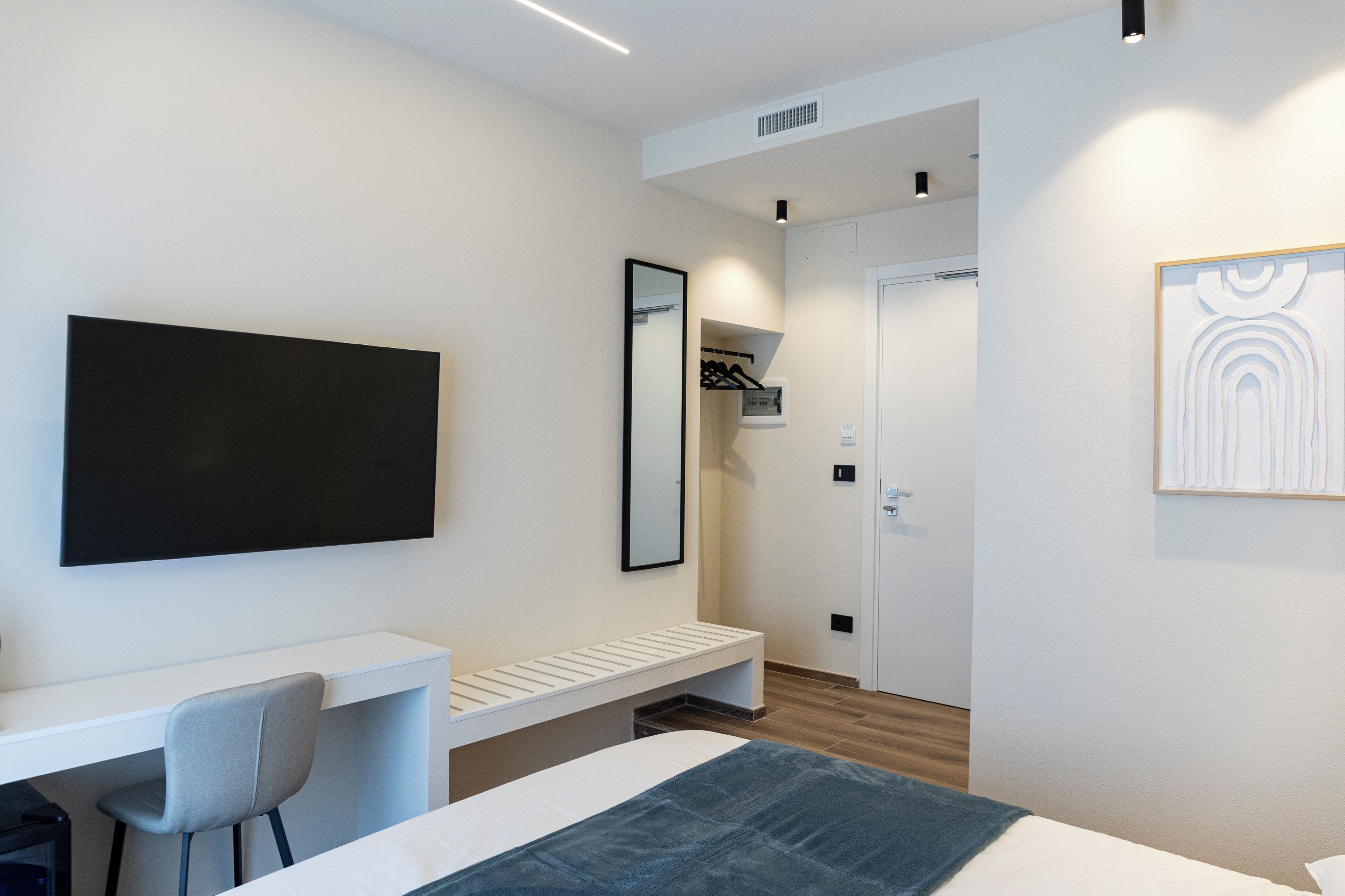 A modern hotel room with a bed, a desk, a chair, a wardrobe, and a flat-screen TV mounted on the wall.