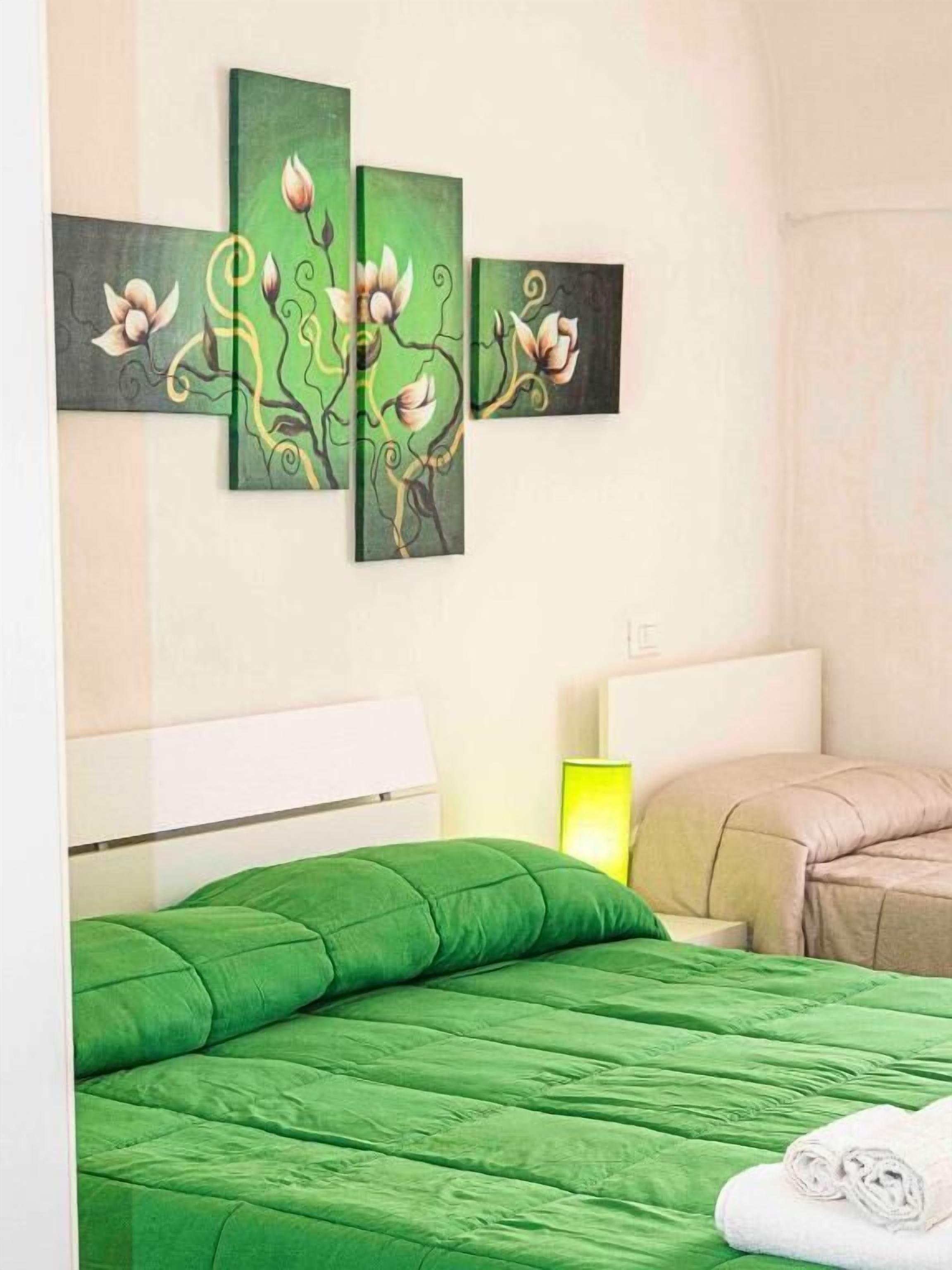 A hotel room with a green bedspread, a white headboard, a wall-mounted painting, a bedside lamp, and a beige sofa.