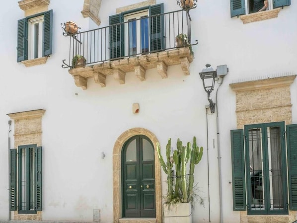 A building with a central arched doorway, green shutters, a balcony with plants, and a cactus.