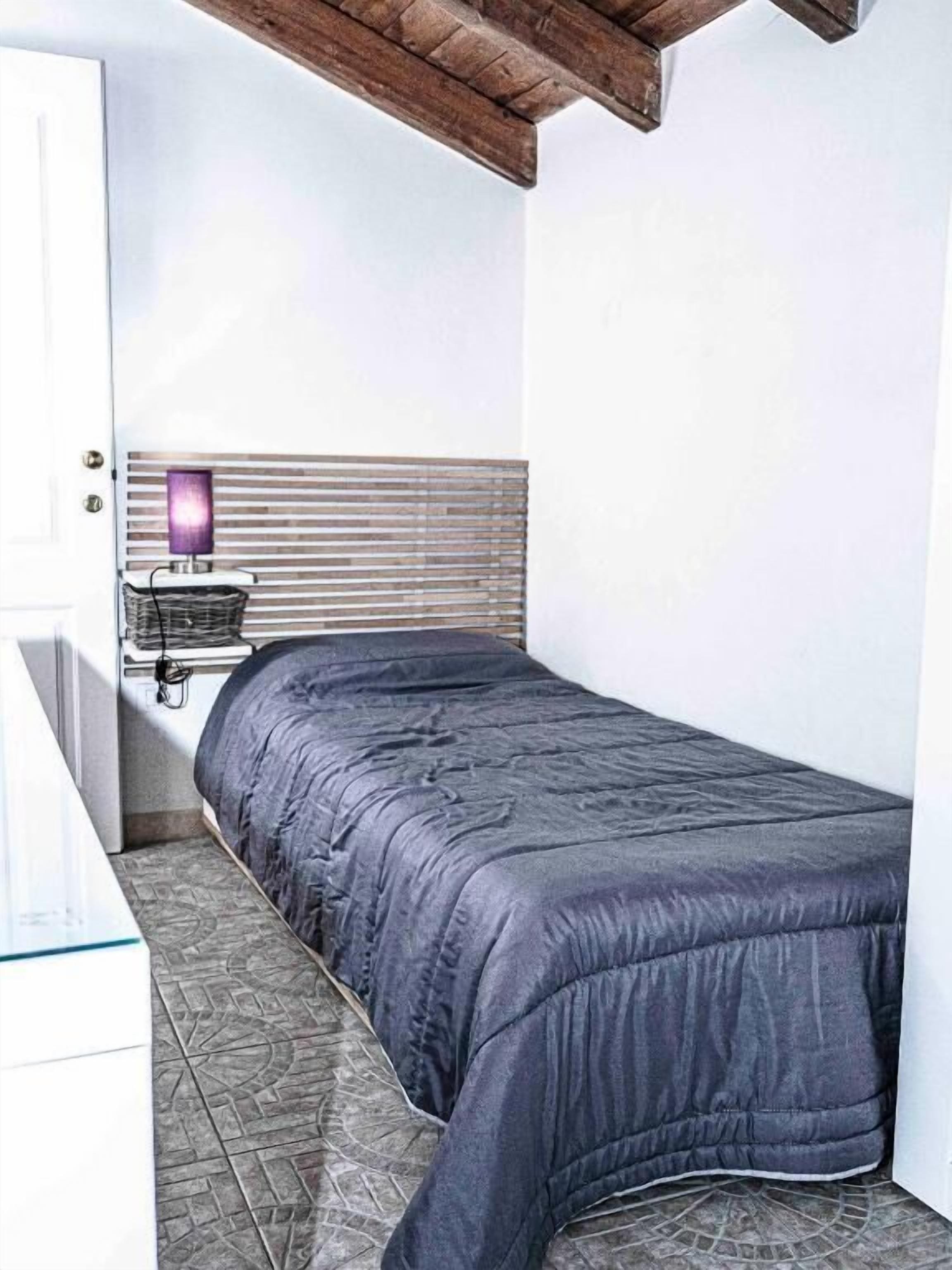 Un lit simple avec une couette de couleur sombre, une table de chevet avec une lampe et une étagère fixée au mur.