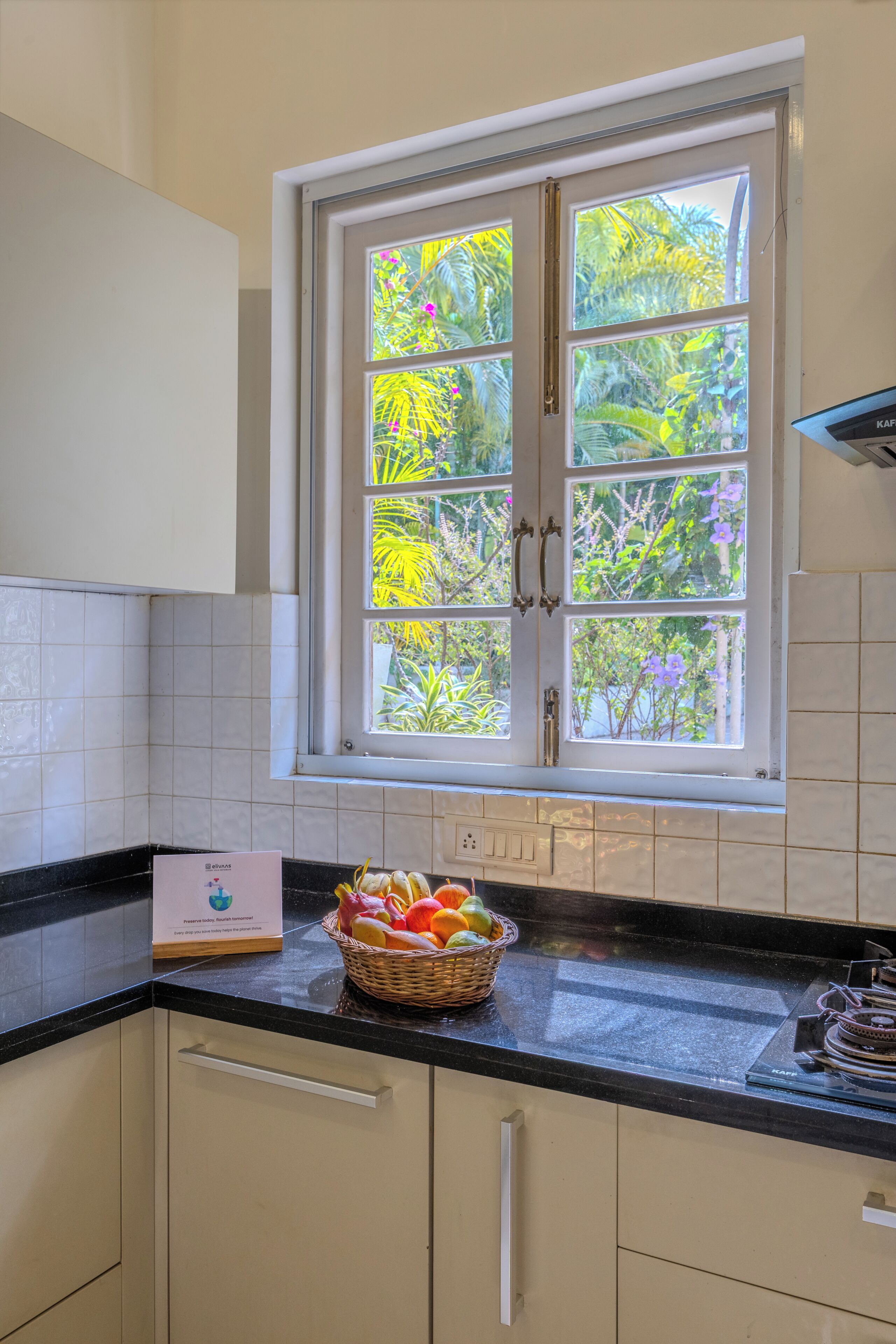 Una cocina con ventana, armarios blancos, encimera negra y cesta de fruta.