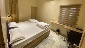 Hotellihuone, jossa on sänky, työpöytä, tuoli, ilmastointilaite, ikkuna, jossa on sälekaihtimet, ja seinälle kiinnitetty hylly.