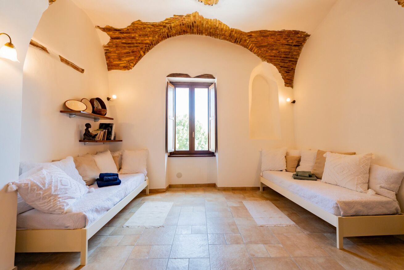 Um quarto com dois sofás brancos, piso de pedra, janela com vista e um arco rústico de tijolos.