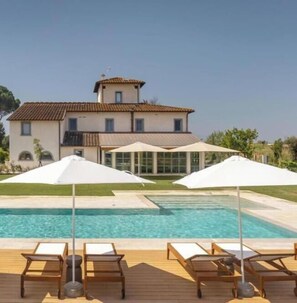 A poolside area with lounge chairs and umbrellas, a pool, and a building with a tiled roof. - Tenuta Asmara Luxury Resort: Executive Suite (Toscana)