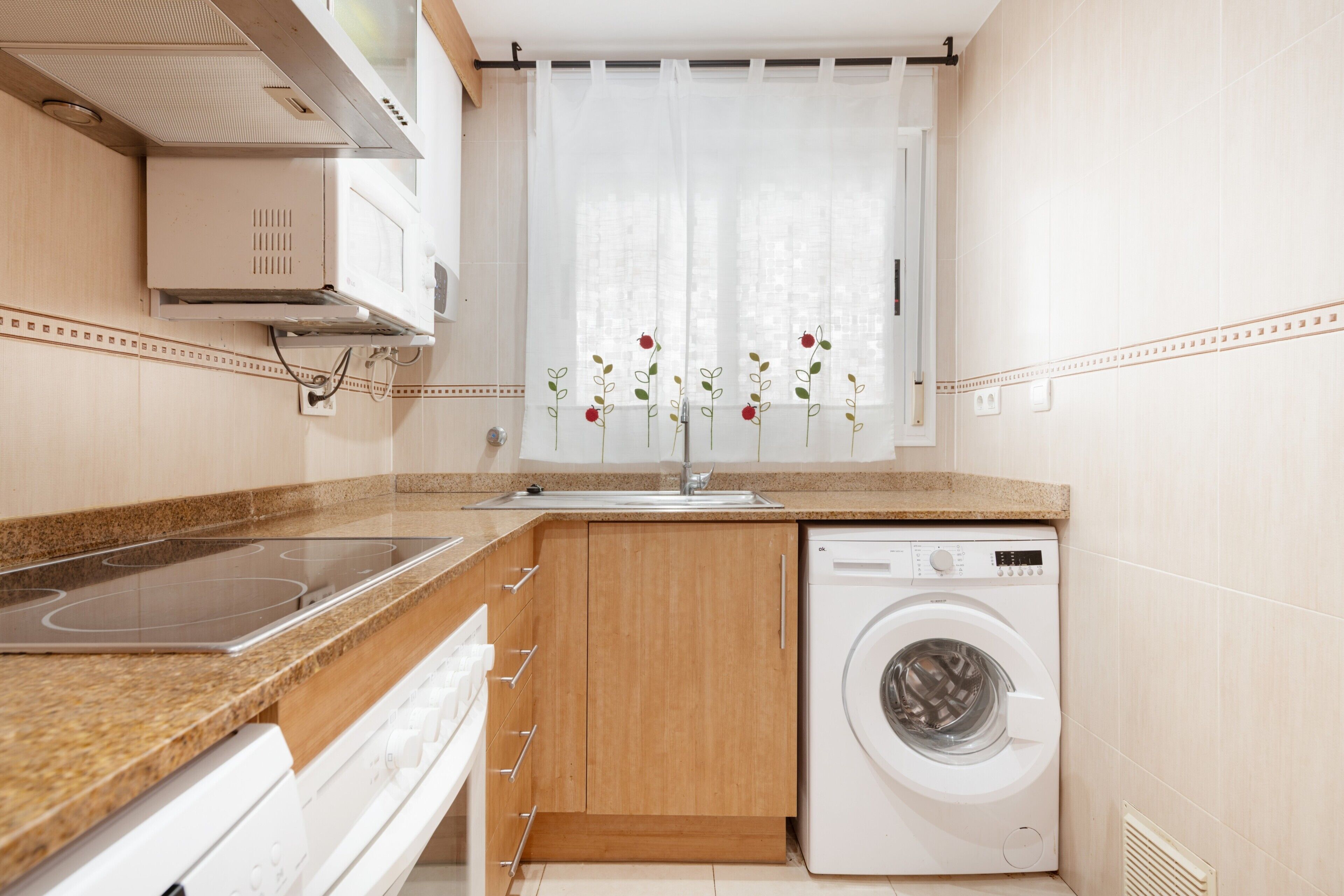 A kitchen with a built-in stove, a washing machine, and a window with a curtain.