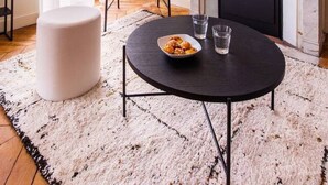 Un salon avec une cheminée, une table ronde sur laquelle se trouve un saladier rempli d’en-cas, un fauteuil en osier et un pouf blanc.