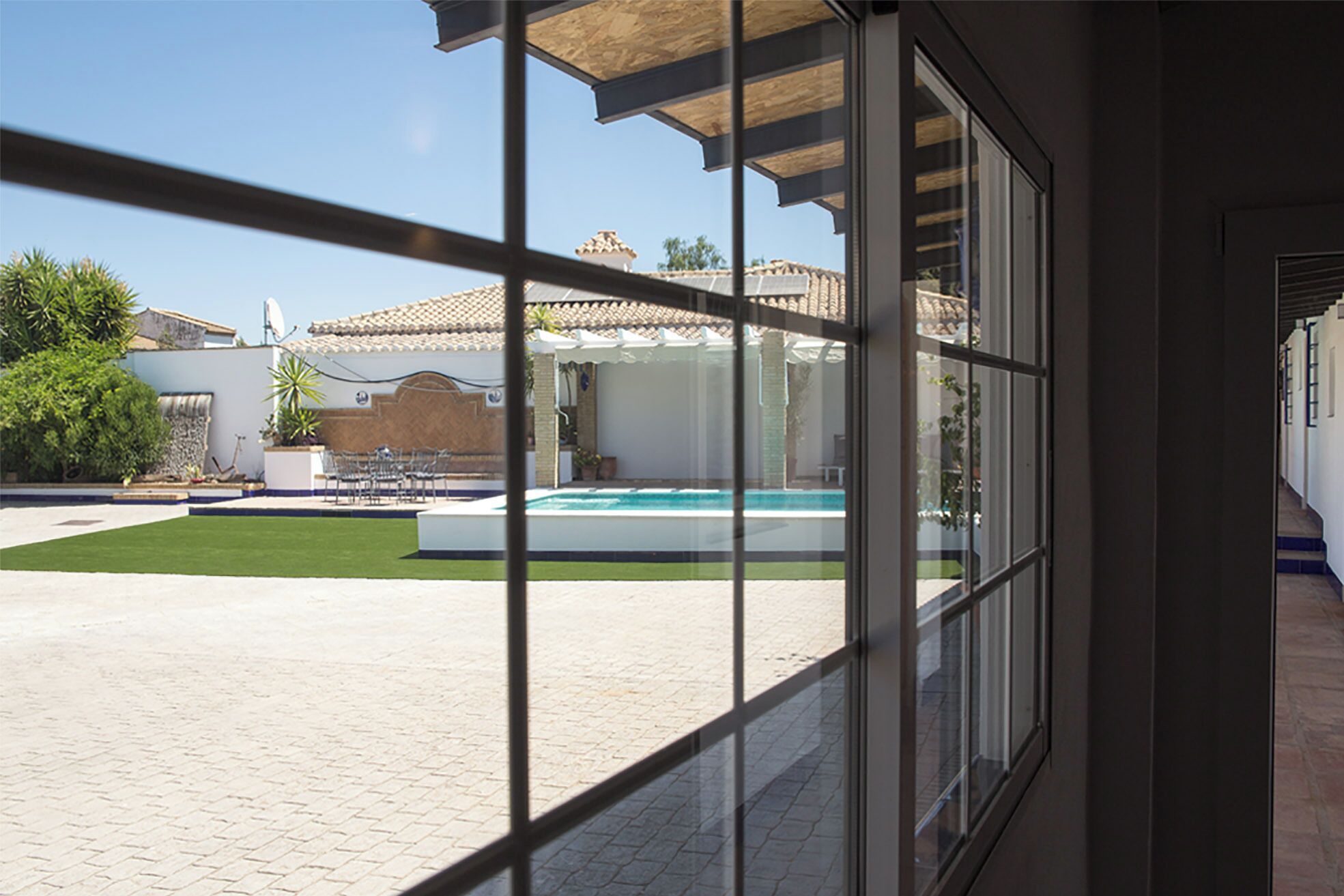 Vista a través de una ventana que muestra una piscina, un patio y un edificio con tejado de baldosas.