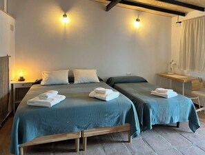 A room with two beds, each with a blue bedspread and white pillows, a bedside table with a lamp, and a small desk with a lamp.