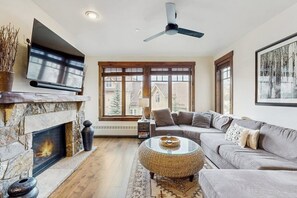 Ein Wohnzimmer mit Kamin aus Stein, einem Flachbildfernseher, Deckenventilator, Sofa und einem großen Fenster.
