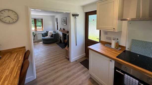 A modern kitchen with a wooden countertop, a built-in stove, and white cabinets. There is a dining area with a wooden table and chairs, and a living room with a sofa and a coffee table.