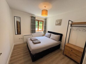 Ein Schlafzimmer mit einem Bett, einem Fenster mit Vorhängen, einem Heizkörper, einer hölzernen Kommode und einem Kleiderständer.