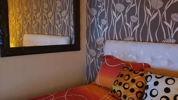 Una cama con un edredón degradado en rojo y naranja, almohadas blancas con diseños negros y anaranjados, un cabecero blanco y una pared con estampado floral.