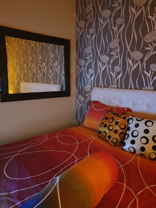 Una cama con un edredón degradado en rojo y naranja, almohadas blancas con diseños negros y anaranjados, un cabecero blanco y una pared con estampado floral.