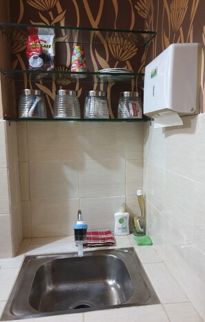 Una cocina con un lavamanos de acero inoxidable, un estante de vidrio con utensilios de cocina y una pared blanca con un diseño decorativo.