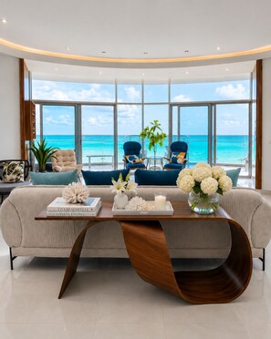 A modern living room with a large window overlooking the ocean, a sofa, two armchairs, a coffee table with decorative items, and a potted plant.