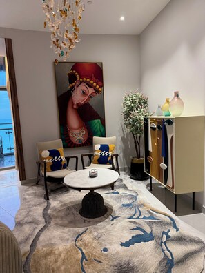 A modern living room with a round table, two armchairs, a large abstract painting, a decorative plant, and a cabinet with vases.