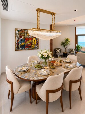 A dining area with a round table, chairs, a chandelier, a painting, and a view of the sea.