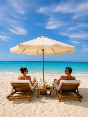 Two people relaxing on wooden lounge chairs under a large umbrella on a sandy beach with clear blue water.