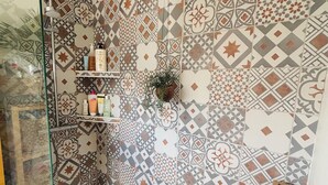 Une salle de bain avec des carreaux décorés, une douche en verre, une baignoire blanche et une étagère en bois où sont rangés les produits de toilette.
