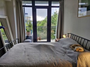 A bedroom with a bed, a dog, a balcony with potted plants, and a view of buildings and trees. - Stylish 1 BDR APT in the heart of Kilmainham (Dublin)