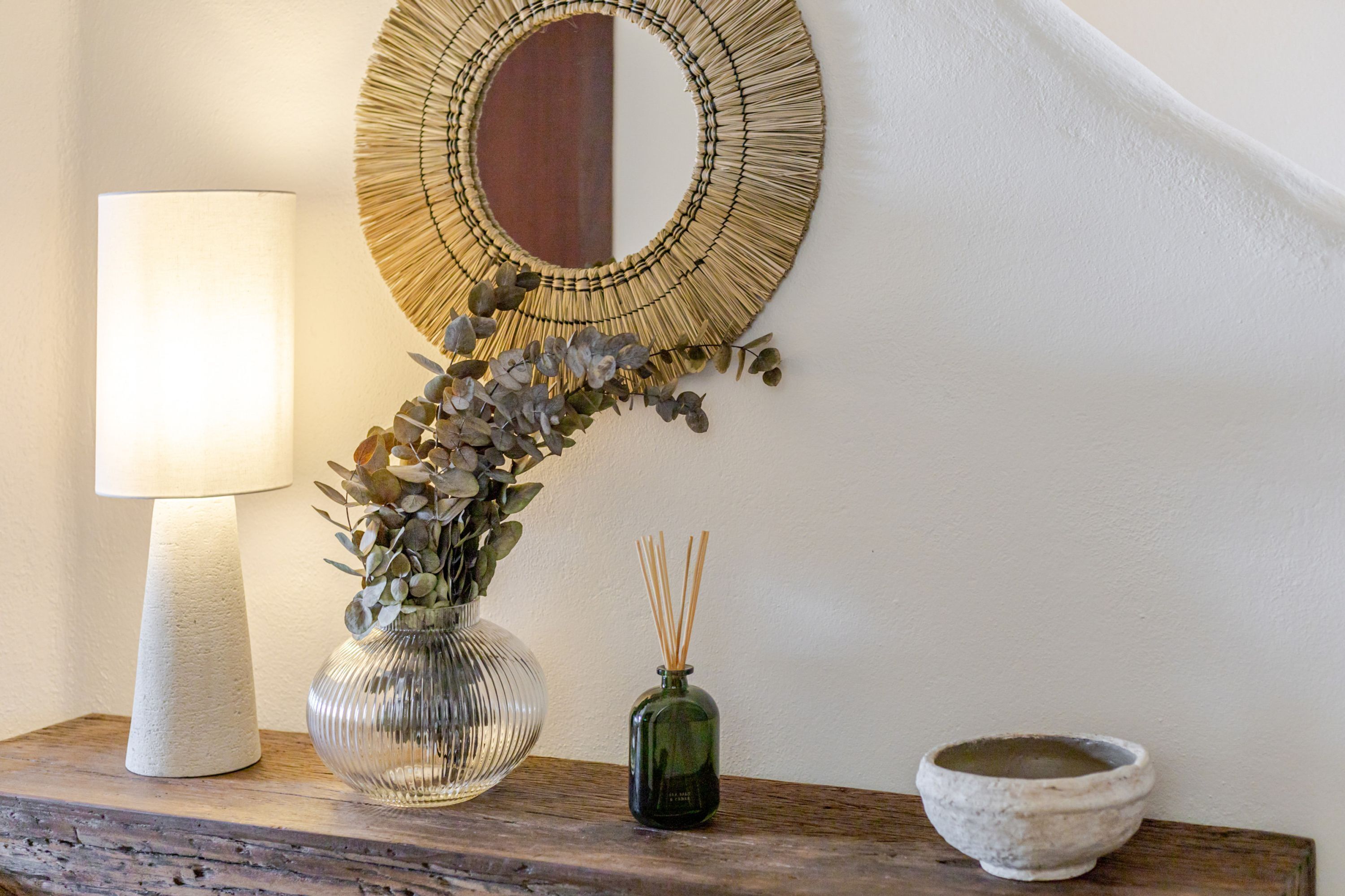 A lamp, a vase with plants, a reed diffuser, and a bowl on a wooden surface.