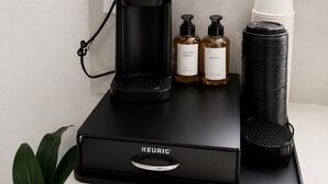 Una cafetera KEURIG sobre una bandeja negra, con dos botellas de café y una pila de vasos de papel.