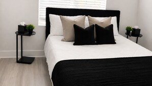 Un dormitorio moderno con una cama, una ventana con persianas, un radiador, una mesita con una planta y un enchufe en la pared.