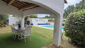 En overdækket terrasse med bord og stole, en lille swimmingpool og en hvid væg med dekorative tallerkener.