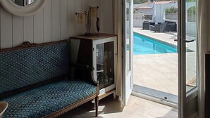 Et værelse med en sofa, et lille skab, et rundt spejl og udsigt til en swimmingpool gennem en åben dør.