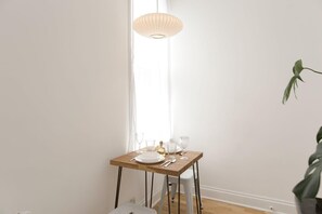 Ein Esszimmertisch ist gedeckt mit Tellern, Gläsern und Besteck, darüber hängt eine Leuchte und in der Ecke steht eine Pflanze.