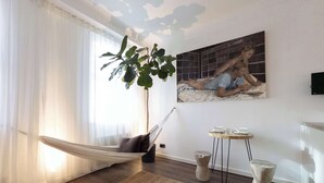 Ein Zimmer mit einer Hängematte, einer großen Pflanze, einem Gemälde, einem kleinen Tisch mit Tassen und einem Fernseher.