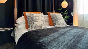 Ein ordentlich bezogenes Bett mit einer dunklen Decke, zwei Kissen mit tropischen Motiven und einem Blumentopf neben dem Fenster.