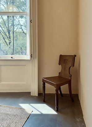 Ein Zimmer mit einem Holzhocker, einem Fenster mit Blick auf Bäume und einer Tür.