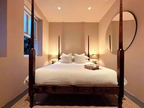 Un lit à baldaquin avec un couvre-lit et des oreillers blancs, deux lampes de chevet, un miroir rond accroché au mur et une fenêtre donnant sur les arbres.