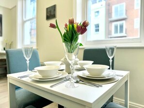 A dining table set with white plates, bowls, cutlery, and a vase with flowers. - Chic Marylebone Village| Premium Mattress & 55”TV (Greater London)