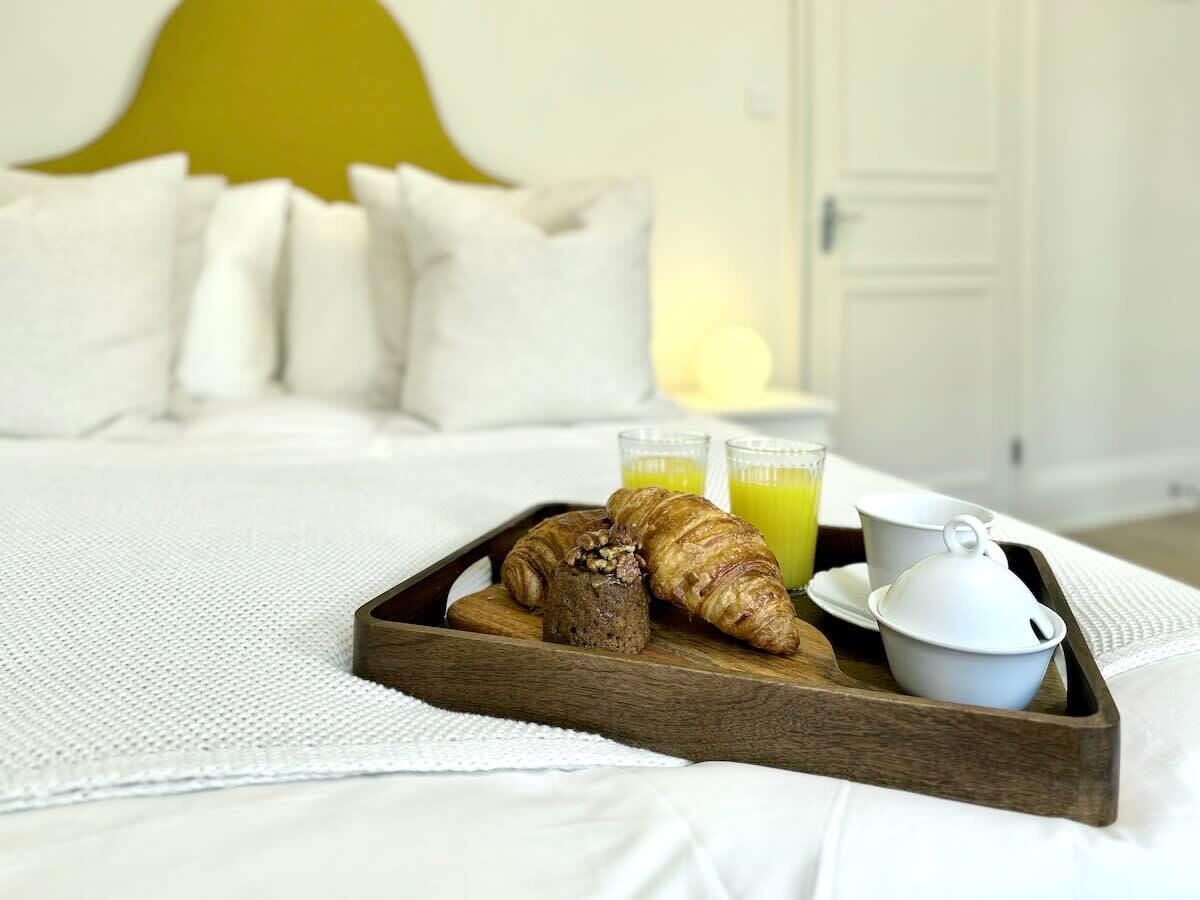 Uma cama com uma bandeja de madeira contendo croissants, um recipiente com geleia e dois copos de suco de laranja.