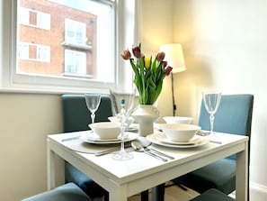 Ein Esstisch ist mit weißen Tellern, Schüsseln und Besteck gedeckt, dazu steht eine Vase mit Tulpen und eine Lampe in einem Raum mit Fenster und Blick auf Gebäude.