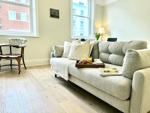 Ein Wohnzimmer mit einer Couch, einem Holzstuhl, einer Platte mit Croissants, einer Zeitschrift und einer Lampe.