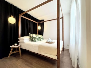 A four-poster bed with a wooden frame, white bedding, and a small bedside table with a plant. - Chic Marble Arch w/ 100" Cinema & Premium Mattress (London)