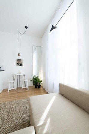 Ein modernes, minimalistisches Wohnzimmer mit einer Couch, einem kleinen Esstisch mit Hocker, einer Topfpflanze und einem großen Fenster mit transparenten Vorhängen.